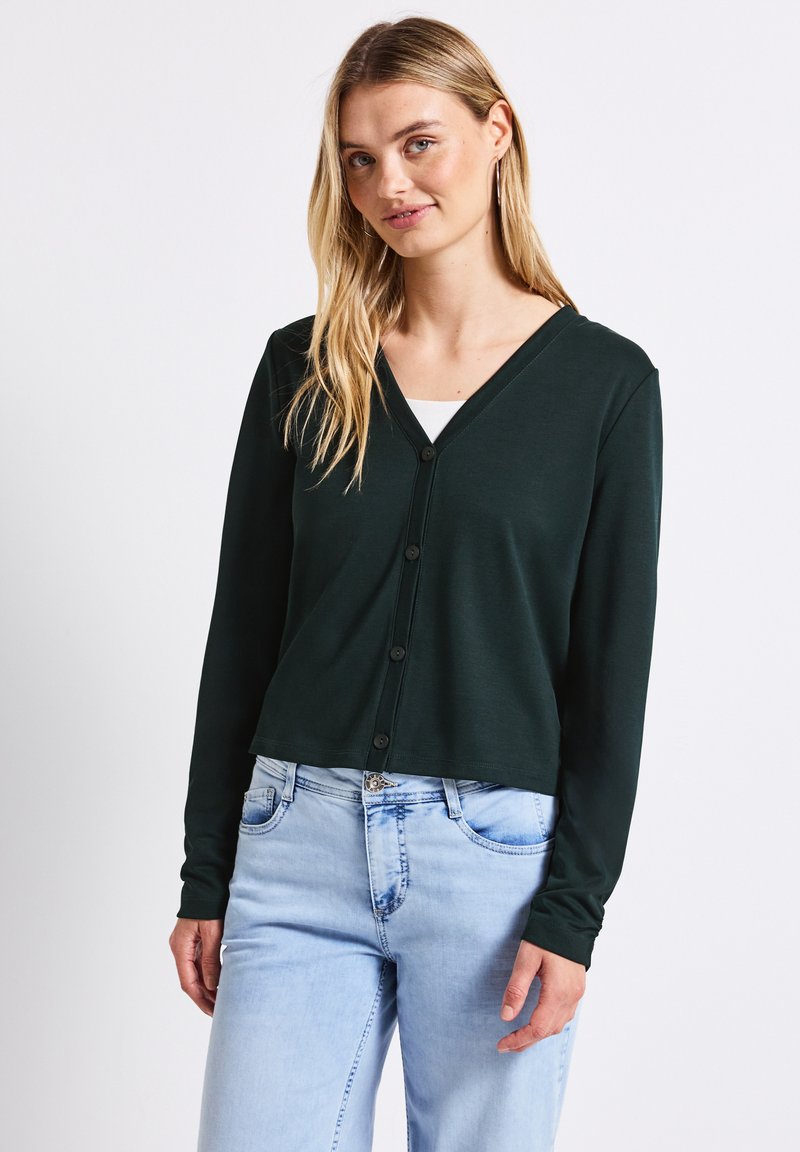 Ciemnozielony krótki kardigan z dekoltem w kształcie litery V, z przodem zapinanym na guziki i długimi rękawami, w połączeniu z jasnoniebieskimi dżinsami. Gładka tekstura.