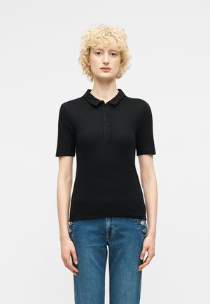 Νεαρό άτομο με κοντά, σγουρά ξανθά μαλλιά, φορώντας μαύρο πόλο μπλουζάκι με κοντά μανίκια και μπλε τζιν με λεπτομέρειες πλευρικών κουμπιών.