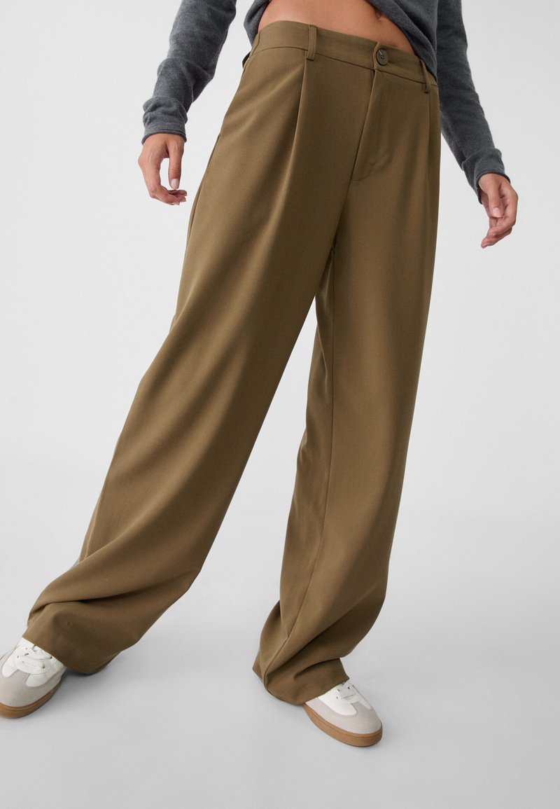 Pantalons larges marron en tissu lisse, avec une fermeture à un bouton et deux poches latérales. Associés à des baskets blanches.