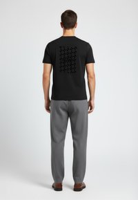 Mann steht mit dem Rücken zur Kamera, trägt ein schwarzes T-Shirt mit dem Aufdruck "Karl Lagerfeld", graue Hosen und braune Schuhe vor einem schlichten Hintergrund.