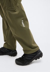 Πόδια με ελαιχρώμα παντελόνια Terrex και μαύρα ορειβατικά παπούτσια σε ανοιχτό γκρι φόντο.