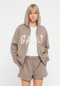 Brown zip-up hoodie with a hood and embroidered "GAP" lettering, paired with matching brown shorts. Fabric appears soft and relaxed.