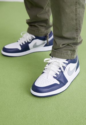 Person iført hvide og marineblå Nike sneakers og olivengrønne bukser står på en lysegrøn gulv.