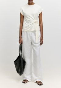 Weißes Kurzarmoberteil mit seitlichem Knoten, kombiniert mit lockeren weißen Hosen. Schwarze Ledertasche und schwarze Sandalen mit minimalen Riemen.