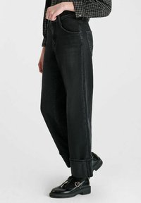 Persoon in hoge zwarte jeans met opgerolde zomen, zwarte enkelsokken, zwarte gespen schoenen en een donker geruit overhemd met lange mouwen.