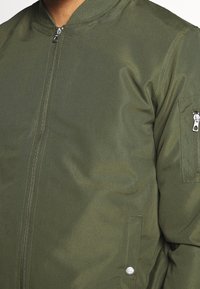 Close-up van een persoon die een olijfgroene bomberjack draagt met een voorrits, een ritszak op de mouw en een drukknopzijzak detail.