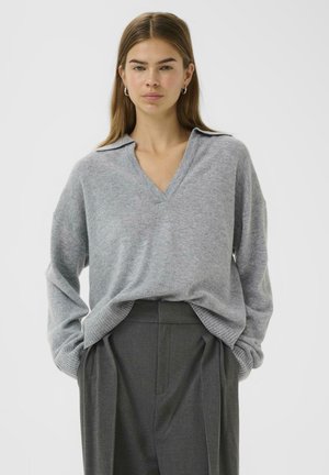Grau gestrickter Pullover mit V-Ausschnitt und Kragen, ausgestattet mit langen, lockeren Ärmeln und geripptem Saum, kombiniert mit hochgeschnittenen, maßgeschneiderten grauen Hosen.