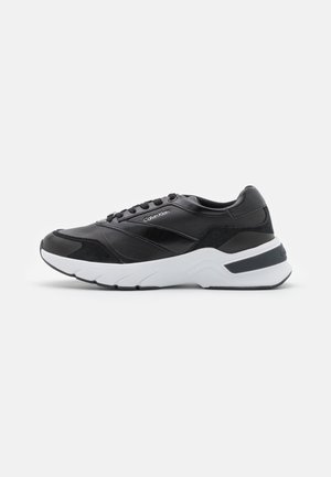 Zapatillas deportivas negras con parte superior de cuero y ante suave, puntera redondeada y suela blanca gruesa con detalles de diseño en capas.