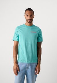 Mand med kort hår og tatoveret arm iført en turkise t-shirt med korte ærmer og logo samt lyseblå jeans, som står mod en hvid baggrund.