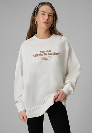Kobieta z długimi włosami nosząca białą bluzę z napisem "Wander With Wonder", czarne legginsy i czarną wzorzystą opaskę na głowę.
