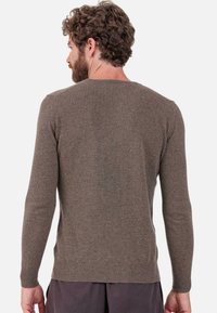 Homme aux cheveux bouclés et à la barbe, portant un pull marron uni à manches longues, vu de dos sur un fond clair uni.