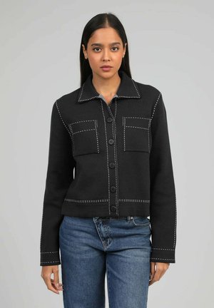 Femme aux cheveux noirs raides portant une veste noire boutonnée avec des surpiqûres blanches et un jean bleu sur un fond gris uni.