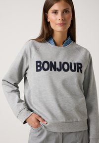 Junge Frau, die einen grauen Pullover mit "BONJOUR" in Marineblau trägt, darunter ein blaues Hemd und eine graue Hose, mit der Hand in der Tasche.