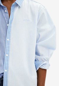 Niebiesko-biała koszula na guziki w paski, z kontrastującymi pionowymi i poziomymi paskami, listwą na guziki i podwiniętymi rękawami.