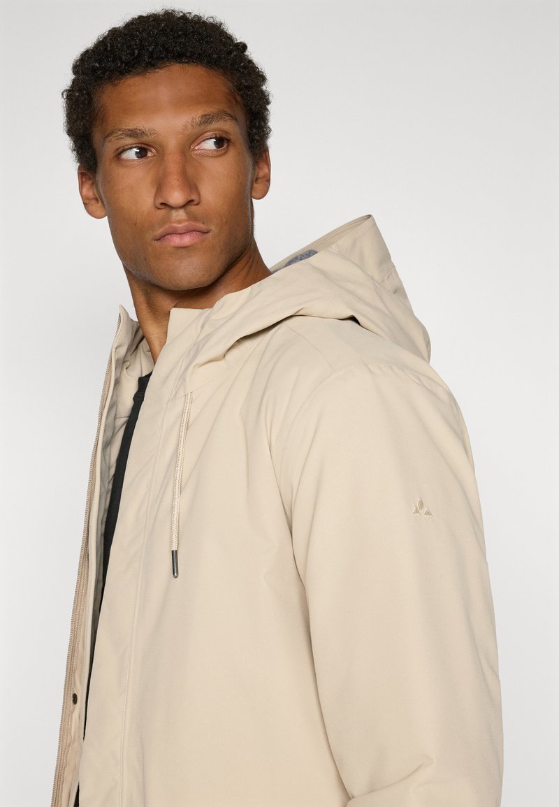Beige hoodie met rits en koord, met een zachte textuur en een logo-accent op de schouder, gedragen over een zwart shirt.