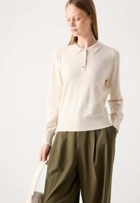 Cremefarvet polo sweater med lange ærmer, krave og placket med tre knapper, båret med olivengrønne, højtaljede bukser.