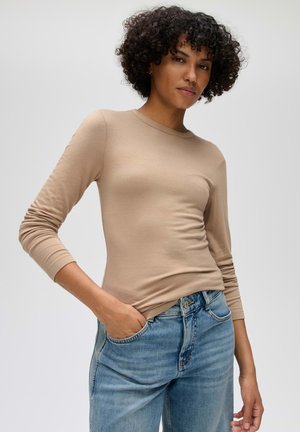 Vrouw met krullend haar, een beige top met lange mouwen en een blauwe spijkerbroek, staand met één hand in haar zak tegen een effen achtergrond.