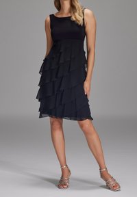 Robe en chiffon noir à plusieurs couches avec un corsage ajusté et des volants. Associée à des sandales argentées à lanières. Texture lisse, design sans manches.