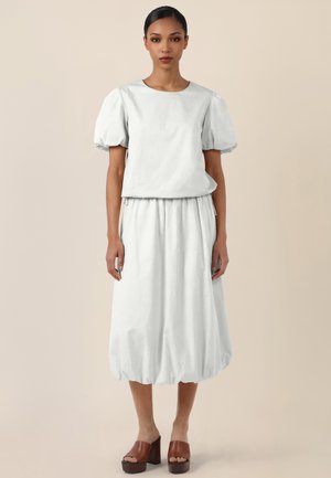 Woman wearing a white puff-sleeve top and matching midi skirt with a gathered hem, paired with brown platform sandals, standing neutral background.
