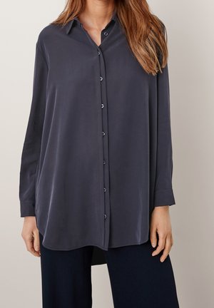 Kobieta ubrana w oversize'ową ciemnoszarą koszulę na guziki z długimi rękawami oraz czarne spodnie na jednolitym jasnym tle.