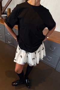 Svart oversize t-shirt, vit tyllkjol med svarta rosettmönster, svart knähöga strumpor och glänsande svarta loafers. Avslappnad lager-på-lager-outfit.