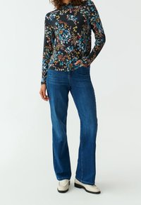 Blumenmuster Langarmshirt aus dunklem Stoff mit blauen, grünen und pinken Blumen. Kombiniert mit blauen Schlagjeans und hellen Stiefeln.