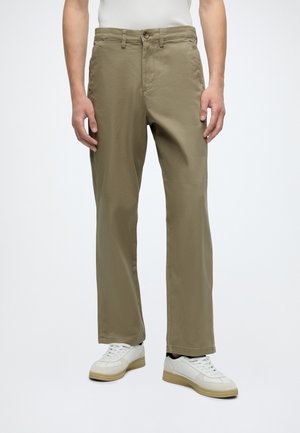 Homme portant un pantalon beige droit et des baskets blanches, debout devant un fond clair uni.