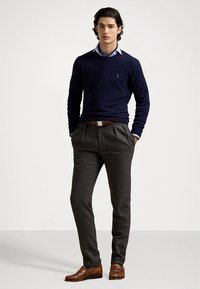 Marinblå tröja över en ljusblå skjorta med krage, grå veckade byxor, bruna läderskor och ett brunt skärp, stående mot en enfärgad bakgrund.