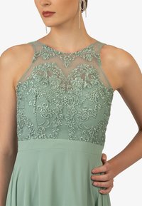 Mintgroene jurk met een doorzichtige geborduurde bovenkant met bloemmotieven en een vloeiende rok. Versierd met delicate pareltjes.