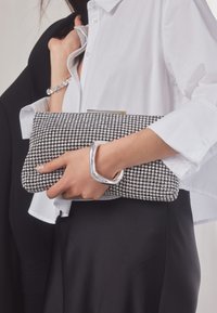 Vrouw in wit overhemd en zwarte rok houdt een fonkelende strass clutch vast met een zilveren armband, terwijl ze een zwart jasje over haar schouder draagt.