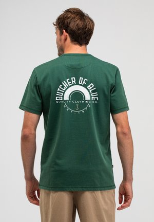Mann trägt grünes T-Shirt mit dem weißen Logo "Butcher of Blue Quality Clothing Co. Est. 1972" auf dem Rücken, kombiniert mit beigen Hosen.