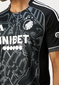 Maillot de football noir avec le blason blanc du lion du FC København, logo du sponsor Unibet et motif de carte stylisé sur le tissu.