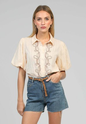 Femme portant un chemisier crème à manches bouffantes et devant brodé, assorti d'une ceinture tressée et d'un short en denim taille haute, debout devant un fond uni.