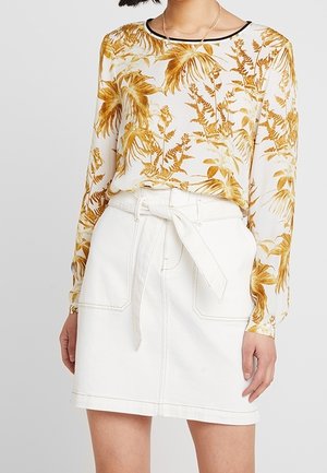 Vrouw draagt een witte rok met een bindceintuur en een witte blouse met lange mouwen en een geel bloemenpatroon.