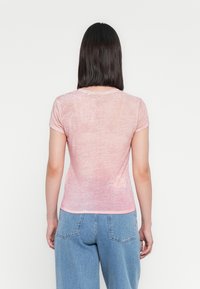 Femeie cu păr drept și negru, purtând un top roz transparent cu mâneci scurte și blugi din denim albastru, întoarsă cu spatele pe un fundal alb simplu.