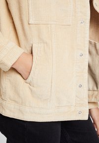 Personne portant une veste en velours côtelé beige avec des boutons-pression, main dans la poche latérale, associée à un pantalon noir.