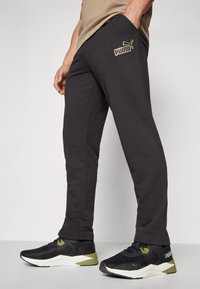 Mörkgrå svettbyxor med en subtil PUMA-logotyp i guld, med sidofickor och en smal bendesign, ihop med svarta sneakers.