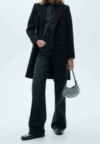 Manteau noir en laine croisé porté sur une chemise boutonnée verte foncée, associé à un jean noir évasé et un petit sac à main texturé.