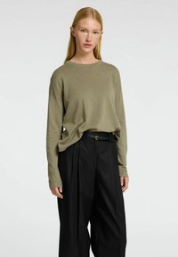 Olivgrüner Langarmpullover aus weichem Material, kombiniert mit hochgeschnittenen, schwarzen, gefalteten Hosen, die eine lockere Passform betonen.