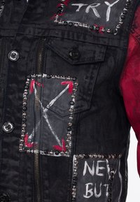 Zwarte denimjack met rode accenten, voorzien van studs, geschilderde graphics en een ritssluiting aan de voorkant. Tekst en pijlontwerpen op de zakken.