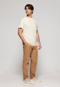 Camiseta de manga corta beige con un pequeño logo, combinada con pantalones marrones claros y zapatillas beige, con un corte relajado y diseño casual.