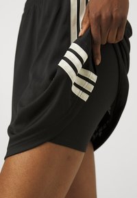 Černé sportovní kraťasy s bílými pruhy po stranách, vyrobené z texturované tkaniny, krátké délky a s mírným rozparkem na lemu.