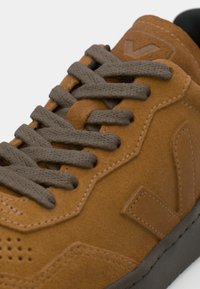 Brown suede sneaker with gray laces, perforated detailing, and a textured rubber sole. Featuring a logo on the heel and side.