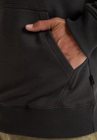 Felpa grigia scura con orlo e polsini a costine, dotata di una tasca frontale e un'etichetta laterale. Tessuto morbido e texture.