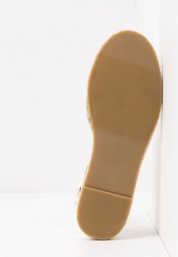 Semelle d'une chaussure plate beige avec une semelle extérieure en caoutchouc texturée positionnée verticalement contre un fond blanc d'angle.