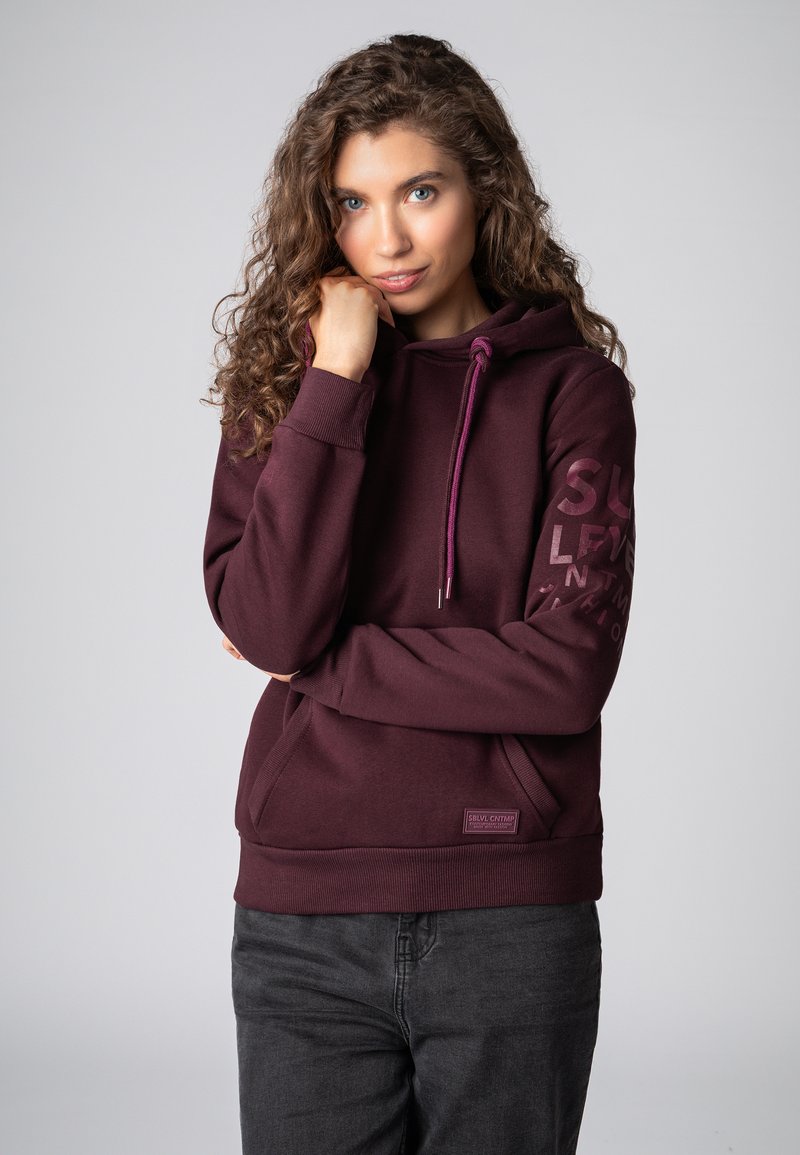 Bordeaux hoodie gemaakt van zacht materiaal, met een voorzak, trekkoordsluiting en bedrukt logo langs de mouw. Textuurlafwerking.