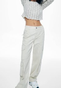 Personne portant un pantalon cargo beige clair, des baskets blanches et bleues, et un pull court gris en maille torsadée, les bras levés.