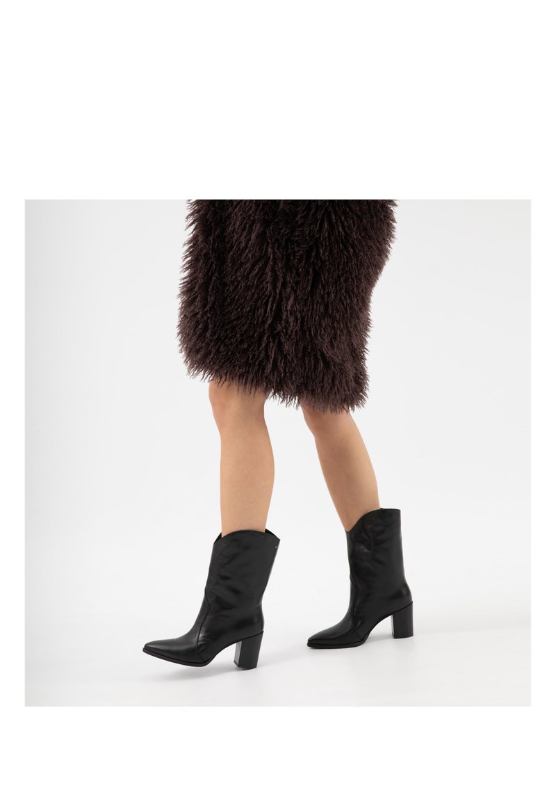 Benenden in zwarte enkellaarzen met hakken en een donkergroene, zachte rok, staand tegen een effen witte achtergrond.