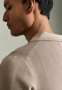 Polo din tricot bej cu modele texturate, guler ribbit și cusături structurale pe umeri, așezat pe un fundal neutru.