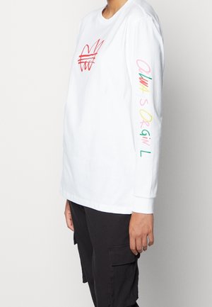 Persoon draagt een wit shirt met lange mouwen, met een rood ontwerp op de borst en meerkleurige tekst op de rechtermouw, gecombineerd met zwarte cargobroek.